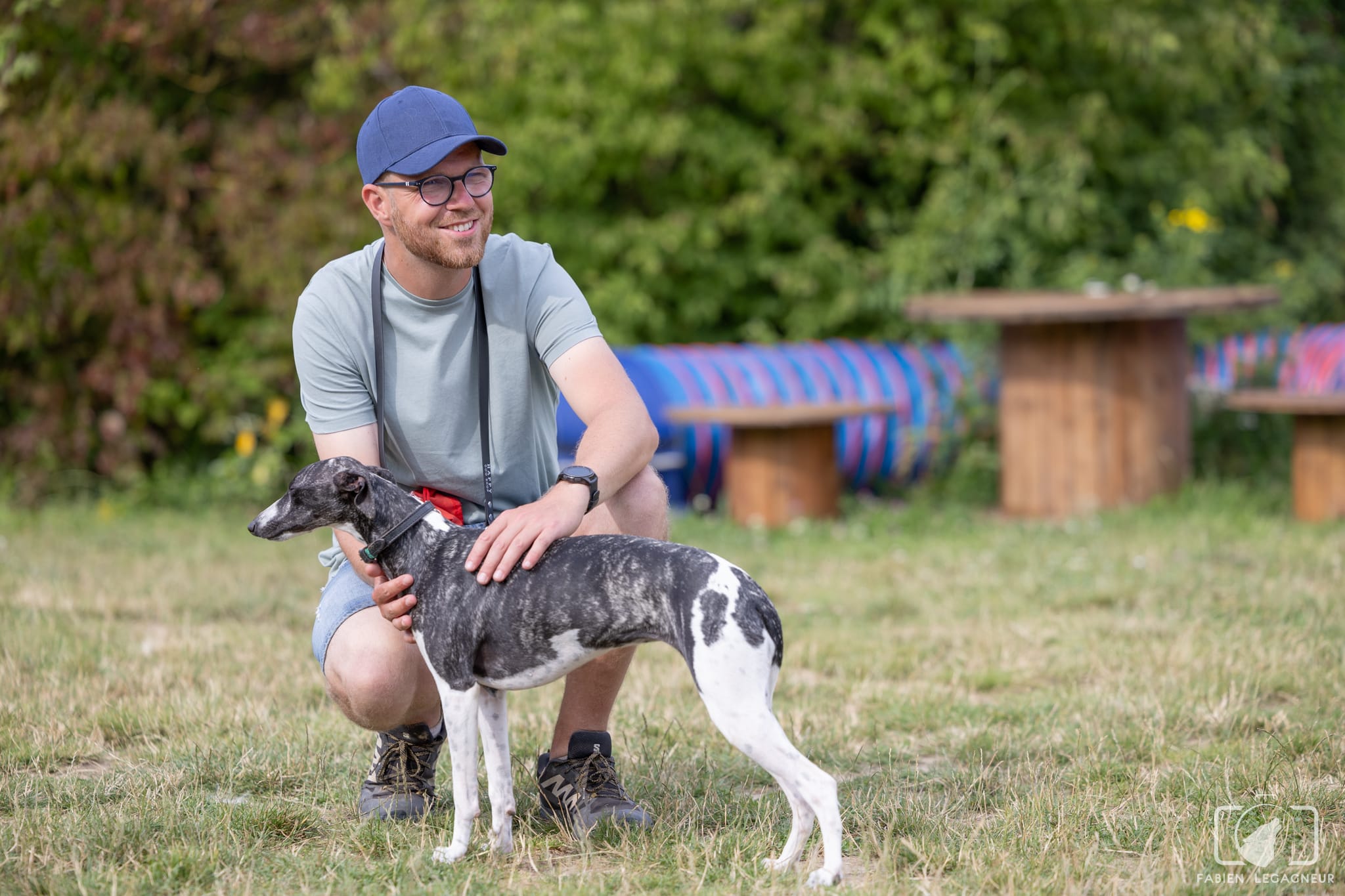 Éducateur canin Pédadog accroupi et complice avec un whippet noir et blanc sur un terrain d'éducation en extérieur